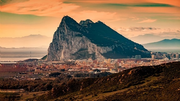 Abendstimmung mit warmen, orangefarbenen Licht: Blick auf Gibraltar – hoher Fels mit kleinem Städchten davor. Wolkenmuster und Berge im Hintergrund.