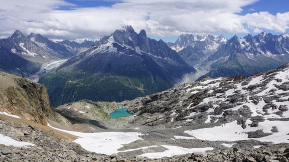 Ansicht leicht von oben: hochalpine Gebirgslandschaft mit zentralem hohen Berg, weitere im Hintergrund, davor teilweise enteistes Gestein und kleiner leuchtend türkisfarbener See. 