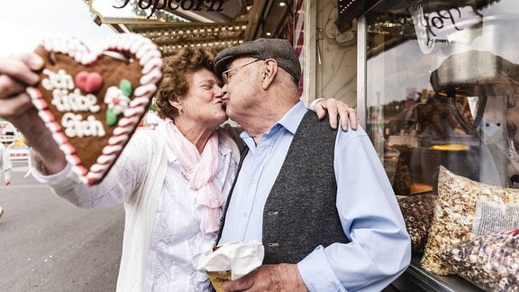 Älteres Paar mit Lebkuchenherz, das sich vergnügte auf dem Jahrmarkt küsst. Aufschrift auf Herz: Ich liebe Dich. Frau streckt Herz Richtung Kamera, dadurch Tiefenunschärfe im Vordergrund.