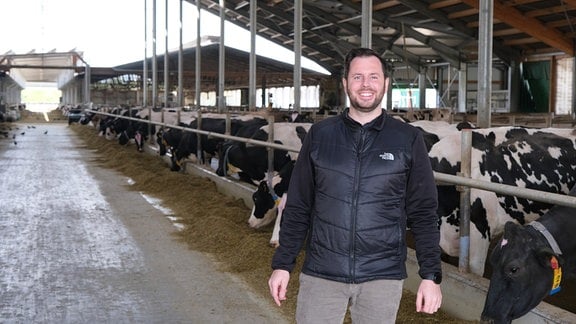 Junger freundlich lachender Mann mit Bart steht vor schwarz-weißen Kühen im halboffenen Kuhstall. Kühe fressen Futter am Boden.