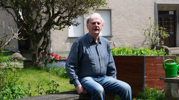 Älterer Mann mit weißen Haaren sitzt neutral-offen-interessiert auf Mauer vor einem Vorgarten, im Hintergrund Baum, Hauswand, Hochbeet, ein paar Tulpen