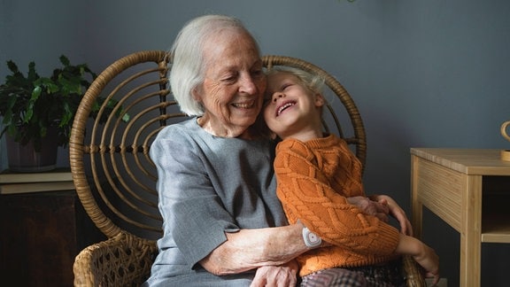 Glückliche ältere Frau mit kinnlangen, eher dünneren Haaren mit lachendem sich ankuschelnden Mädchen auf Schoß