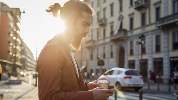 Seitenaufnahme: Mann mit Bart und kleinem Zopf schaut mit Kaffeebecher in Hand auf Handy, Gegenlicht durch Sonne, im Hintergrund Altbau-Fassade