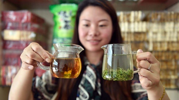 Eine Frau hält zwei Glastassen nebeneinander, in der einen Tasse grüner Tee, in der anderen schwarzer.