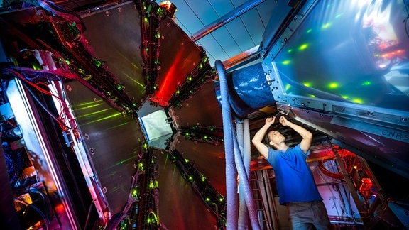 Der HADES-Detektor von Innen: Das Innere einer technischen Anlage mit vielen Leuchten und Kabeln. Ein Mann im blauen T-Shirt schraubt etwas daran.
