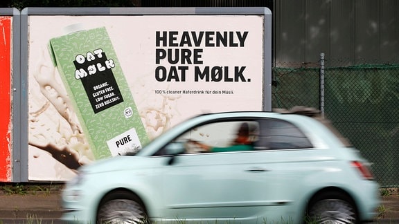 Werbeplakat mit Hafermilch-Packung und Schriftzug englisch "Heavily Pure Oatmølk" – hundert Prozent cleaner Haferdrink für dein Müsli. Und vorbeifahrender neuer Fiat 500 mit Bewegungsunschärfe