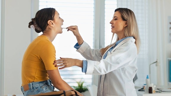 Eine Ärztin untersucht die Halsschmerzen eines jungen Patienten und schaut mit einem hölzernen Zungenspatel in den Hals.