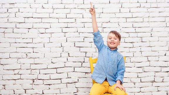 Ein Schüler mit blauem Hemd und gelber Hose sitzt auf einem Stuhl vor einer weißen Ziegelwand und meldet sich. Gestreckte Körperhaltung, Kopf nach oben gerichtet, freudiges Gesicht.