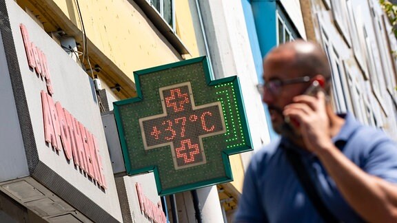 Typische Temperaturanzeige an Apotheke in grünem Kreuz mit 37 Grad, davor unascharf Mann mit Handy am Ohr, Blick leicht von unten