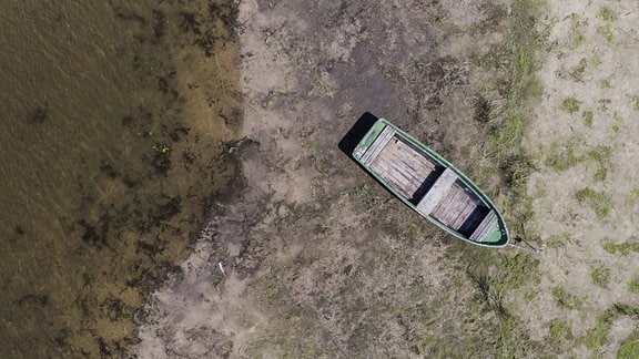 Ansicht von Oben: Leeres Holzboot auf trocknem, erdigen Untergrund. Links etwas Wasser, rechts sandig mit Grasbüscheln.