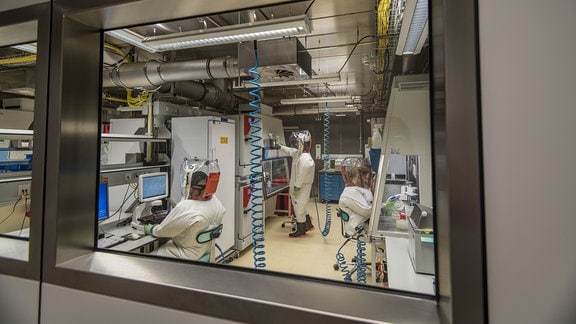 Hinter einer Scheibe arbeiten mehrere Personen in Vollschutzanzügen in einem Labor
