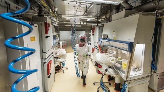 Ein Mann läuft in einem Vollschutzanzug durch ein Labor, zwei weitere sitzen an Arbeitsplätzen