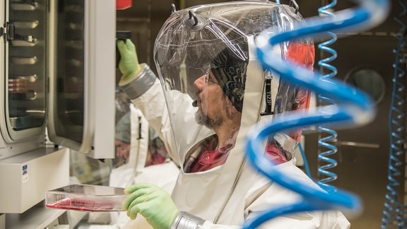Ein Mann in einem Vollschutzanzug in einem Labor hält eine Probe in der Hand und öffnet die Tür eines Geräts