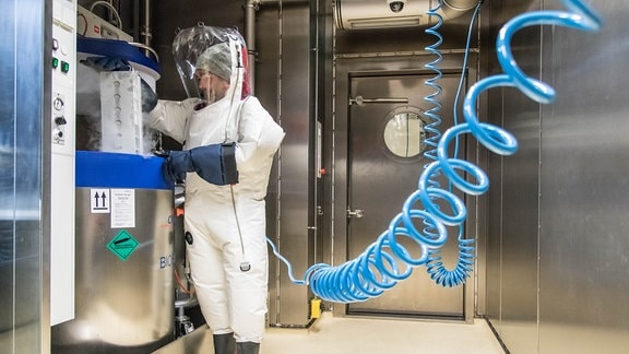 Ein Mann in einem Vollschutzanzug, an dem ein blaues Kabel zur Versorgung mit Luft hängt, nimmt etwas aus einem sogenannten Cryotank