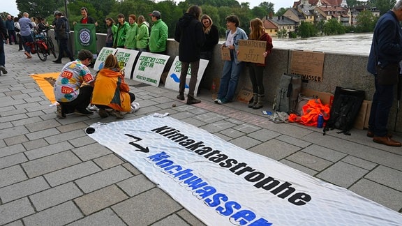 Kleine Protestansammlung auf Brücke über Fluss mit Hochwasser. Transparent am Boden mit Aufschrift "Klimakatastrophe Hochwasser"