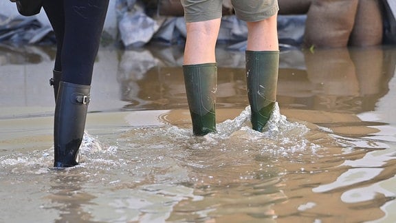 Ausschnitt Beine und Gummistiefel von zwei Personen, die durch Hochwasser schreiten, im Hintergrund Sandsäcke