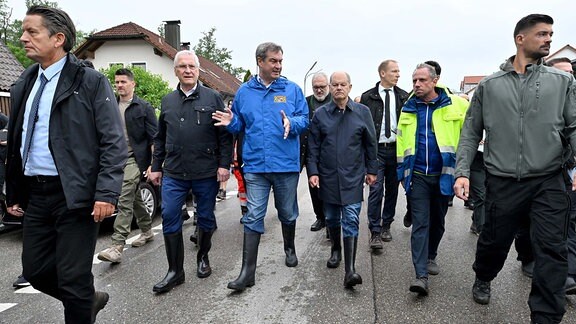 Joachim Herrmann, Markus Söder, Olaf Scholz tragen bei einer Ortsbesichtigung Gummistiefel in einer Gruppe anderer Männer. Die anderen Männer tragen normale Schuhe.