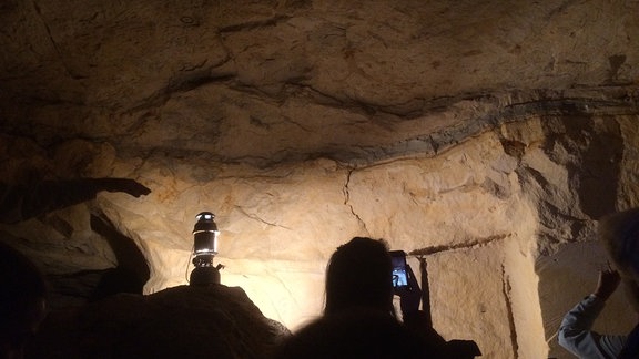 Innenaufnahme eine Höhlenwand unter Beleuchtung mit dunkleren Gesteinschichten, die sich deutlich vom Rest der Wand unterscheiden, im Vordergrund eine Lampe und eine Person, die ein Foto macht.