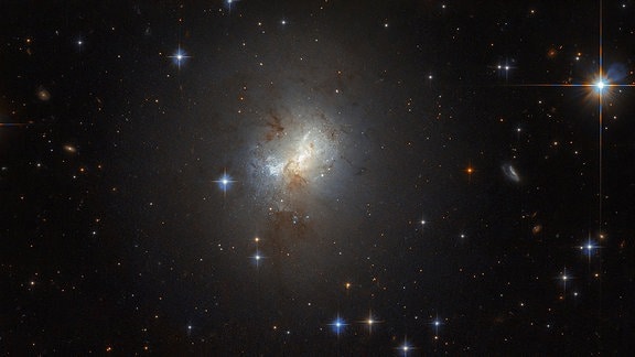 Fotografie des Weltraumteleskops Hubble von der Galaxie ESO 495-21. Zu sehen ist ein heller Fleck mit dunklen Wolken darin, drum herum befinden sich verschiedene helle Sterne im Vordergrund.