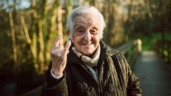 Ältere Frau mit Hörner-Geste mit der rechten Hand lacht in die Kamera, Außenaufnahme, im Hintergrund Natur