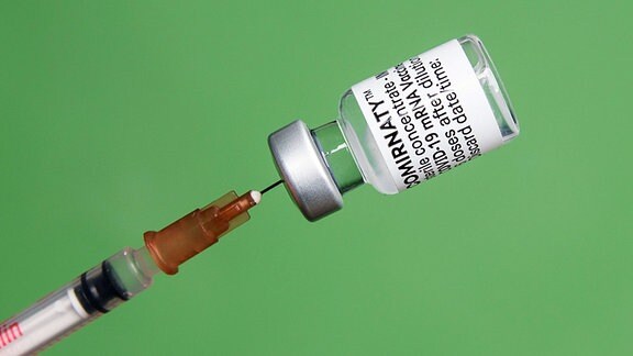 Vor einem grünem Hintergrund, wird mit Hilfe einer Impfspritze eine Impfdosis mit dem mRNA-Impfstoff Comirnaty, BNT162b2, Biontech / Pfizer aus einer Ampulle gezogen