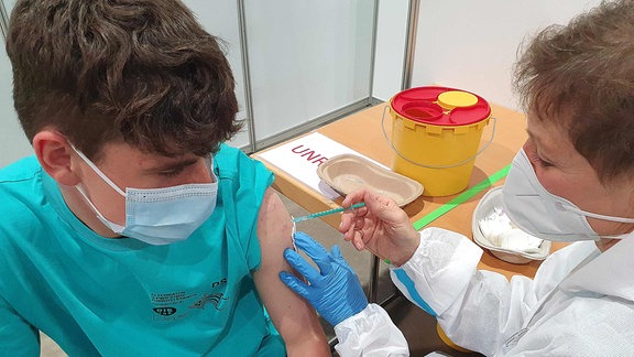 Männlicher Jugendlicher mit Maske bekommt von einer medizinischen Mitarbeiterin in Schutzkleidung eine Impfung. Ansicht von schräg oben.