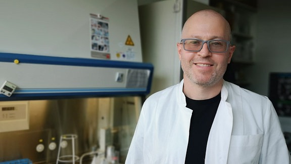 Porträtaufnahme eines Mannes mit Glatze, einer Brille mit grauem Rahmen und einem weißen Laborkittel, der in einem Labor steht. 