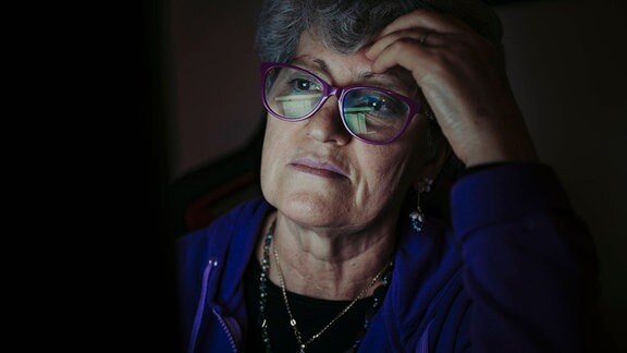 Ältere Frau mit Brille und grauen Haaren sitzt auf Hand aufgestützt im Dunkeln, in Gesicht und Brille spiegelt sich ein Bildschirm, in den sie konzentriert blickt, Hintergrund schwarz