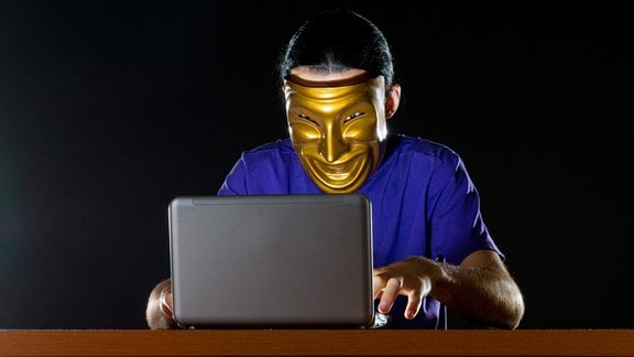 Ein Mann sitzt mit einer Maske vor einem Laptop.