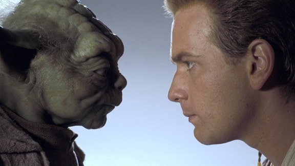 Zwei Personen blicken sich an. Links Yoda mit typisch außerirdischem Aussehen, wulstiges Gesicht, spitze Ohren und rechts Obi-Wan Kenobi mit Aussehen eines jungen Mannes.