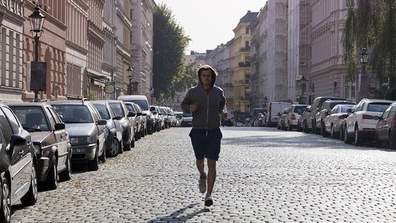 Junger Mann joggt auf Kopfsteinpflaster mittig Richtung Kamera, links und rechts Gründerzeithäuser und parkende Autos