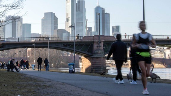 Frau joggt am Mainufer in Frankfurt, weitere Personen beim Spazieren, im Hintergrund eine Brücke und die Skyline mit vielen Wolkenkratzern