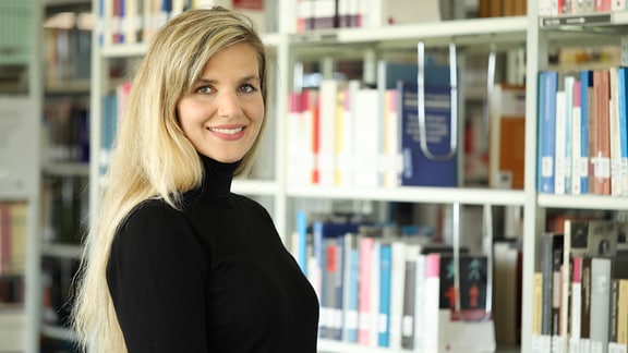 Lächelnde Frau mit langen blonden Haaren und schwarzem Rollkragenpullover im Portrait, Bücherregal unscharf im Hintergrund.