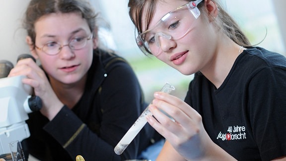 Zwei Schülerinnen bei einem Laborexperiment. Eine trägt Schutzbrille und hält ein Reagenzglas, beide betrachten es. Im Vordergrund Pipette und Proben in Gläsern mit Deckel.