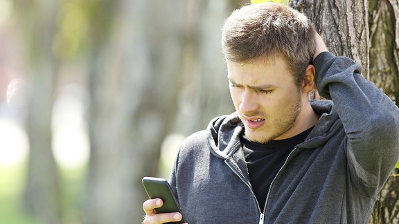 Ein junger Mann mit blonden Haaren sieht auf sein Smartphone und hat einen verwirrten Gesichtsausdruck, an einem Baum stehend, Hintergrund unscharf