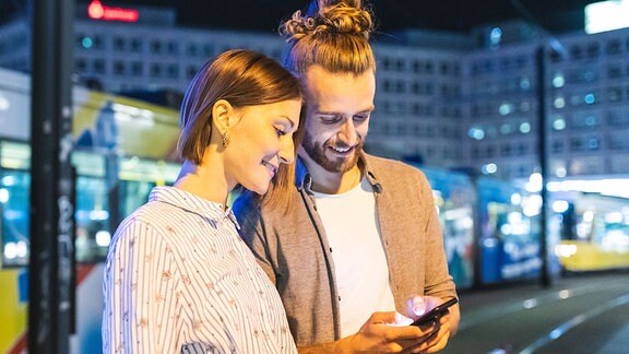 Ein junges Paar ist abends in einer Stadt unterwegs; beide sehen auf ein Telefon
