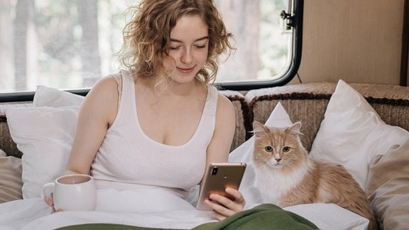 Frau mit schulterlangen, gewellten Haaren sitzt in einem Wohnwagen in Bett mit Kaffeetasse und Handy, neben ihr eine Katze. Frau schaut auf Handy, Katze in Kamera.