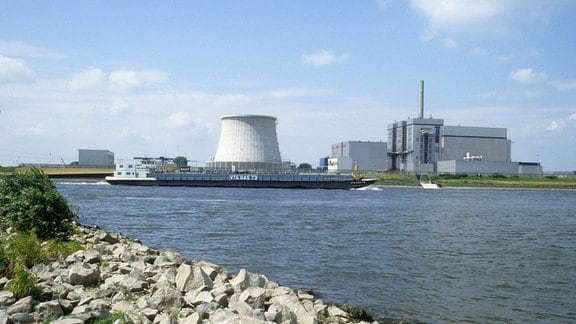 Kernkraftwerk Kalkar: Großer, flacher Kühlturm, daneben fabrikähnliches Gebäude, im Vordergrund Fluss und Steine, sonnig