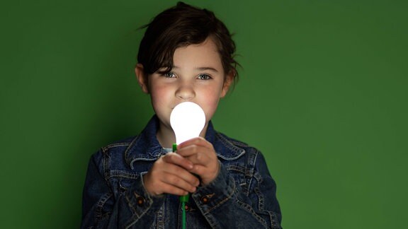 Kind in grünem T-Shirt, das in einem dunklen Raum steht und eine Glühbirne nahe am Gesicht hält.
