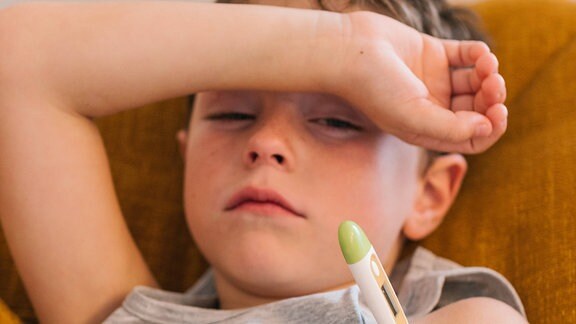 Erschöpft aussehendes Kind liegt mit Fieberthermometer unter einem Arm auf einer Couch.