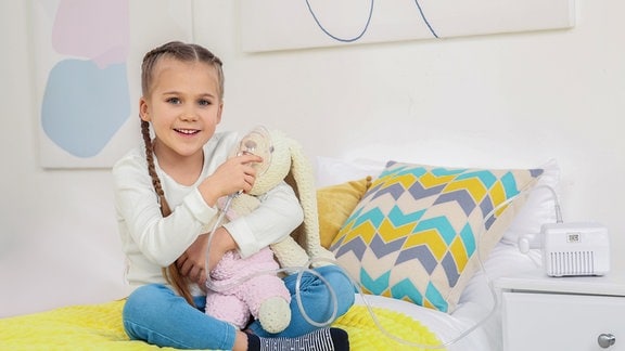 Kleines Mädchen sitzt mit Spielzeughase und Vernebler zur Inhalation zu Hause im Bett