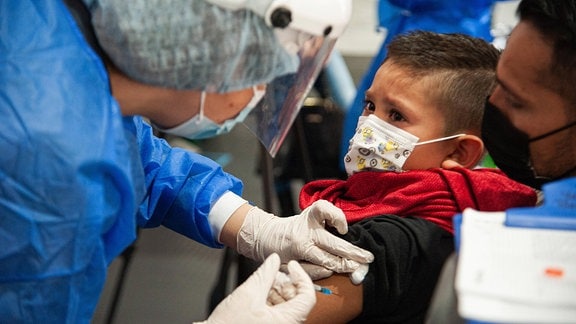 Ein Impfarzt in blauem Kittel, mit Mundschutz impft einen kleinen Jungen, der auf dem Schoß seines Vaters sitzt, eine Maske trägt und den Arzt etwas ängstlich anschaut.