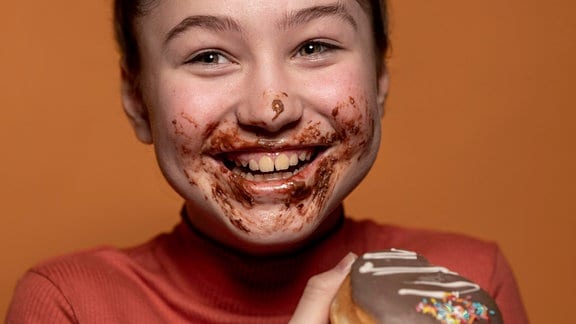Lachendes Mädchen mit schokoverschmiertem Mund hält Schoko-Donut ohne Loch