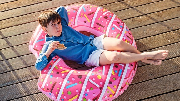 Ein Junge sitzt in einem aufblasbaren Donut mit einem Brötchen in der Hand