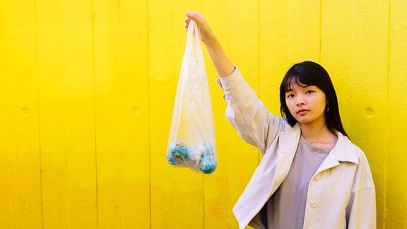 Junge Frau vor gelber Wand hält mit neutralem Blick und leicht geneigtem Kopf eine Plastiktüte mit drei kleinen Erdkugeln hoch.