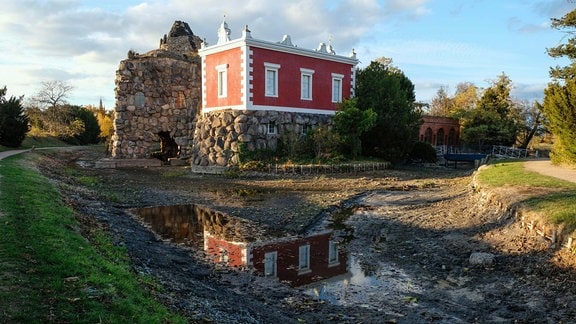 Blick auf einen ausgetrockneten Teich, im Hintergrund ein rotes Haus
