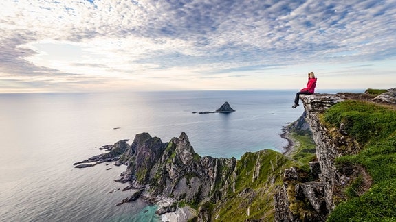 Weibliche Person mit roter Outdoor-Jacke sitzt auf einer Klippe bzw. Felsvorsprung in Norwegen, niedrigere Felsen ragen bis ins Meer, Moosbedeckte Felsen, tiefe Sonne hinter Schleierwolken
