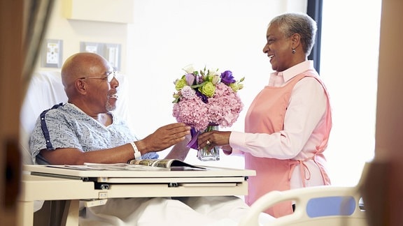 Eine Frau überreicht einem Mann im Krankenbett einen Blumenstrauß in einer Vase.