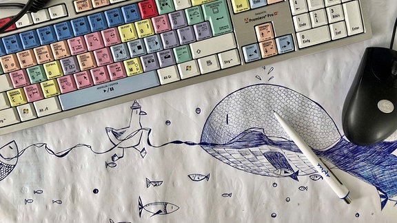 Arbeitplatz mit Tastatur, Computermaus, Köpfhörern und Stift. Darunter ein goßes Blatt Papier, auf das Fische und eine Möwe gezeichnet wurden.