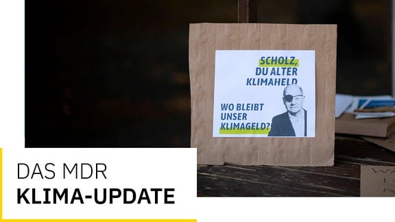 Plakat-Pappe für Demonstration mit Olaf Scholz mit Augenklappe und Spruch Scholz, du alter Klimaheld, wo bleibt unser Klimageld. Und Text Das MDR Klima-Update.